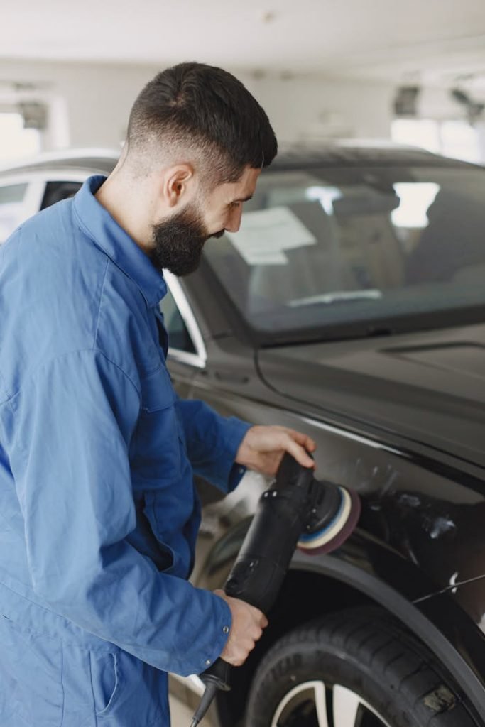 Mechanic wearing blue uniform polishing car body with machine in workshop.